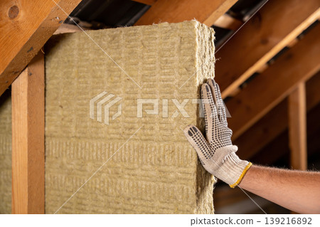 Worker installing thermal insulation material in attic space with wooden beams 139216892