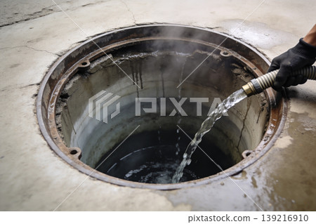 Pouring water into an open manhole with steam rising from the dark depths below Pouring water into an open manhole with steam rising from the dark depths below 139216910