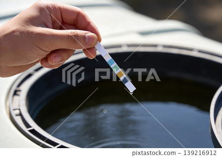 Hand holding a water quality test strip over a large container of murky water Hand holding a water quality test strip over a large container of murky water 139216912
