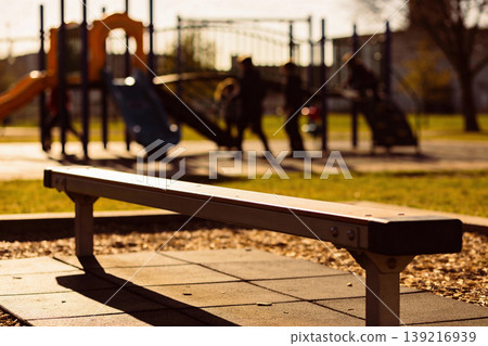 Golden hour sunlight casts long shadows on a playground bench with children playing in the background Golden hour sunlight casts long shadows on a playground bench with children playing in the background 139216939