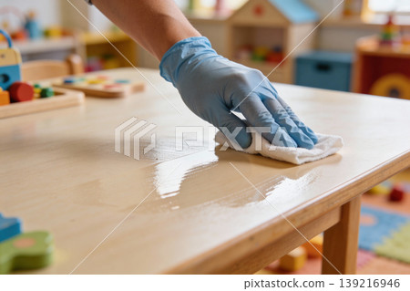 Hand in blue glove cleaning a wooden table with a white cloth in a classroom Hand in blue glove cleaning a wooden table with a white cloth in a classroom 139216946