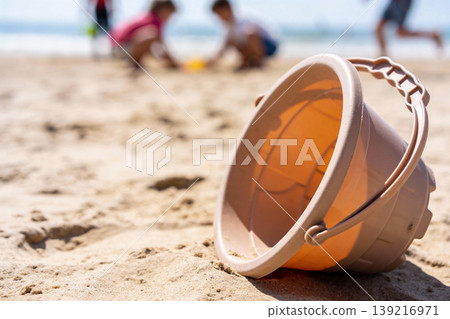 Plastic beach bucket resting on sand with children playing in the background Plastic beach bucket resting on sand with children playing in the background 139216971