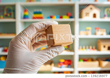 Hand wearing protective glove holding a wooden block in a playroom with toys on shelves Hand wearing protective glove holding a wooden block in a playroom with toys on shelves 139216977