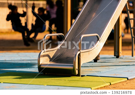 Children playing on swings and slide at a sunny playground during golden hour Children playing on swings and slide at a sunny playground during golden hour 139216978