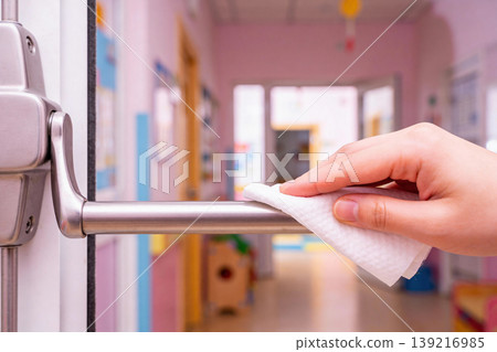 Close-up of a hand wiping a metal door handle with a white wet wipe in a brightly lit hallway 139216985