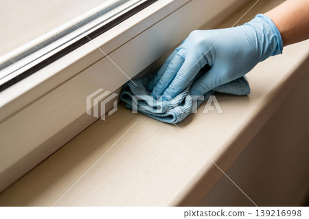 Hand wearing blue glove wiping a window sill with a microfiber cloth for cleaning Hand wearing blue glove wiping a window sill with a microfiber cloth for cleaning 139216998