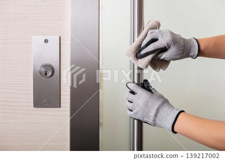 Hands wearing gloves and cleaning a metal door handle with a cloth for hygiene 139217002