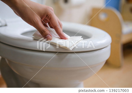 Close-up of a hand wiping down a white toilet seat with a wet wipe for hygiene 139217003