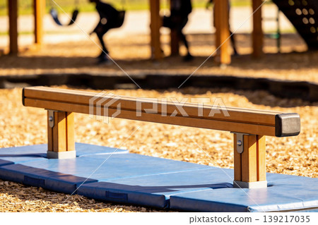 Wooden balance beam on a playground with soft mats and blurred children playing in the background 139217035