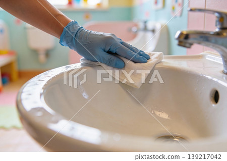 Hand wearing blue glove wipes down a white sink with a cloth for hygiene and cleanliness 139217042