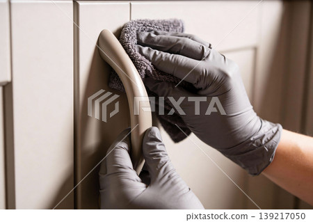 Hands wearing gray gloves cleaning a cabinet handle with a microfiber cloth 139217050
