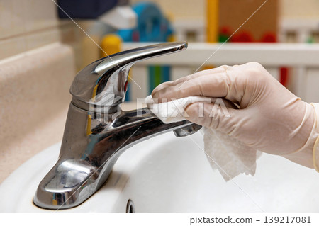 Hand wearing glove wipes down a chrome faucet with a wet wipe for hygiene 139217081