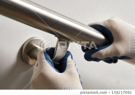 Hands in protective gloves installing a stainless steel grab bar on a white wall 139217092