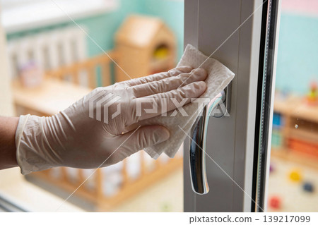 Gloved hand wipes down a window handle in a brightly lit room with toys in the background Gloved hand wipes down a window handle in a brightly lit room with toys in the background 139217099