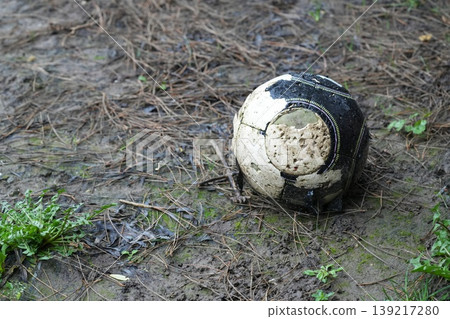 A worn-out and muddy soccer ball rests forgotten on the wet, earthy ground amidst pine needles A worn-out and muddy soccer ball rests forgotten on the wet, earthy ground amidst pine needles 139217280