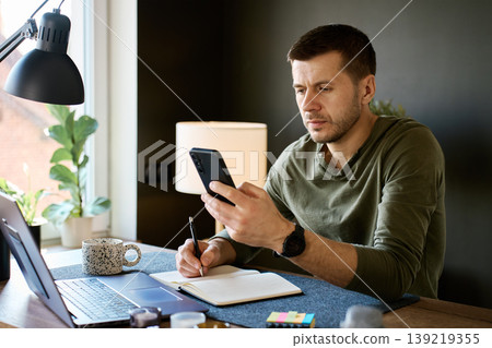 Man using smartphone and writing notes at home desk Man using smartphone and writing notes at home desk 139219355