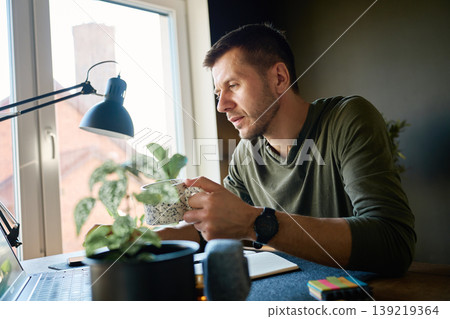 Man with coffee working on laptop by window at home Man with coffee working on laptop by window at home 139219364