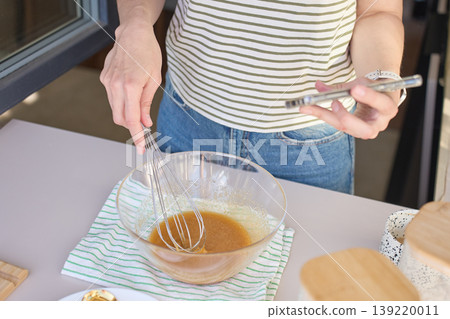 Woman mixes batter with whisk in glass bowl while holding smartphone Woman mixes batter with whisk in glass bowl while holding smartphone 139220011