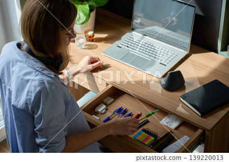 Woman arranging stationery in open desk drawer near laptop in home office Woman arranging stationery in open desk drawer near laptop in home office 139220013