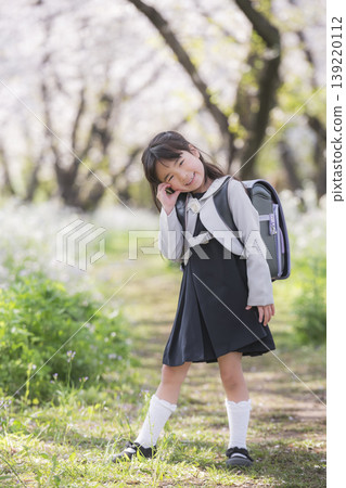 A young girl, a first-grader in her school uniform, smiles amidst a row of cherry trees. A young girl, a first-grader in her school uniform, smiles amidst a row of cherry trees. 139220112