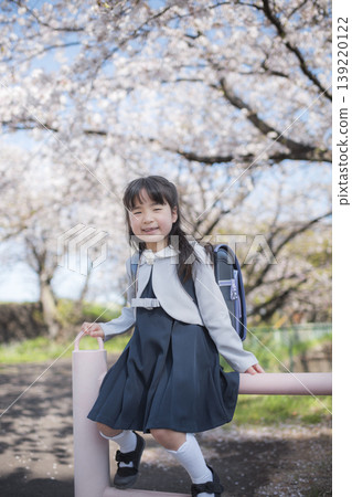 一位小學生小女孩坐在那裡微笑，背景是盛開的櫻花。 139220122