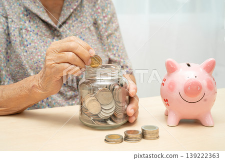 Asian elderly woman putting coin into pink piggy bank for saving money and insurance, poverty, financial problem in retirement. 139222363