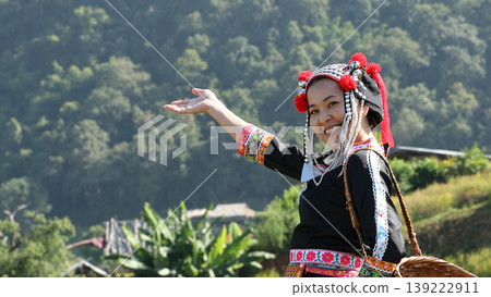Smiling ethnic woman in traditional tribal dress gesturing with hand in rural village, showing cultural pride, hospitality, and joyful lifestyle in scenic nature 139222911