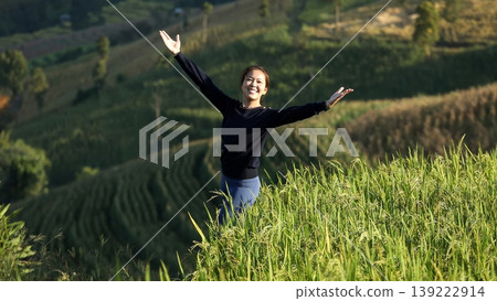 Happy Asian woman standing in green rice field on mountain, turning and raising arms with joy, celebrating nature, freedom, and healthy rural lifestyle in peaceful environment 139222914