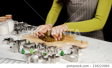 Close-up of hands kneading dark green cookie dough on wooden board, surrounded by metal Christmas cookie cutters and festive tree decorations 139223060