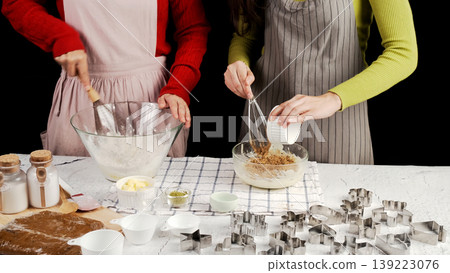 Two people preparing cookie dough together, adding ingredients and whisking in glass bowls surrounded by baking tools and cutters, capturing a warm homemade baking moment in a cozy kitchen Two people preparing cookie dough together, adding ingredients and whisking in glass bowls surrounded by baking tools and cutters, capturing a warm homemade baking moment in a cozy kitchen 139223076