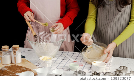 Two women baking cookies together as one whisks flour, other mixes cookie dough. Cozy kitchen scene with baking tools, matcha powder, and butter on counter 139223077