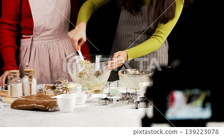 Two women stir cookie dough in glass mixing bowl on kitchen table with baking tools and ingredients, captured during homemade cookie preparation session Two women stir cookie dough in glass mixing bowl on kitchen table with baking tools and ingredients, captured during homemade cookie preparation session 139223078