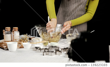 Close-up of hands mixing cookie dough in glass bowl, surrounded by baking tools and cookie cutters, with camera capturing baking process behind scenes 139223103