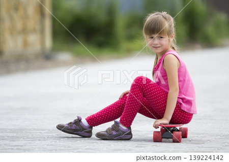 Pretty young long-haired blond child girl in casual pink clothing sitting on skateboard on paved suburb street on blurred sunny summer green background. Children activities, games and fun concept. Pretty young long-haired blond child girl in casual pink clothing sitting on skateboard on paved suburb street on blurred sunny summer green background. Children activities, games and fun concept. 139224142