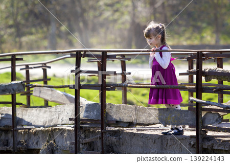 Pretty small blond long haired girl in nice red dress stands alone on old cement bridge leaning on wooden railings looking intently down on warm sunny day. Children curiosity and fantasies concept. Pretty small blond long haired girl in nice red dress stands alone on old cement bridge leaning on wooden railings looking intently down on warm sunny day. Children curiosity and fantasies concept. 139224143