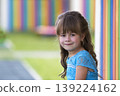 Portrait of little fashionable blond girl in blue dress, with gray eyes and beautiful long hair smiling in camera on bright blurred colorful background. Beauty and innocence of childhood concept. 139224162