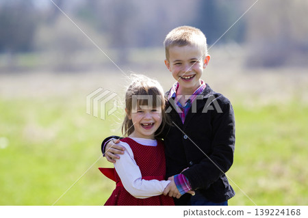 Portrait of beautiful blond smiling children with funny child teeth in smart clothes standing together outdoor on blurred background, older brother embracing sister. Happy careless childhood concept. 139224168