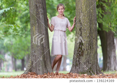 Portrait of a young woman standing near big tree trunk in summer park outdoors. 139224169