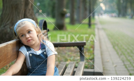 Little serious child girl sitting alone on a bench in summer park. Little serious child girl sitting alone on a bench in summer park. 139224259