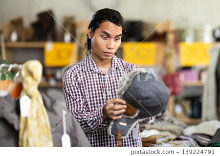 In chain shop, amazed Indonesian man choose hat cap with earflaps In chain shop, amazed Indonesian man choose hat cap with earflaps 139224791