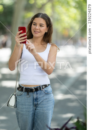 Girl with phone walks along sunlit street, photographs architectural objects Girl with phone walks along sunlit street, photographs architectural objects 139224879