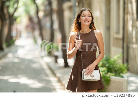 Cute young girl wearing stylish short summer dress with white handbag on shoulder walking along shady tree lined street enjoying spring day in Barcelona 139224880