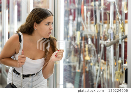 Interested young girl looking at crafted swords in souvenir shop Interested young girl looking at crafted swords in souvenir shop 139224906