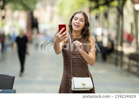 Beautiful stylish young girl in dress with white handbag walking and posing on city street on summer day and taking selfies with mobile phone 139224929