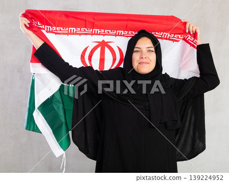 Balanced smiling Muslim woman in traditional black hijab holds flag of Iran 139224952