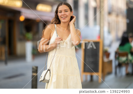 Cheerful girl in white dress walking along the street among architecture with bag 139224993