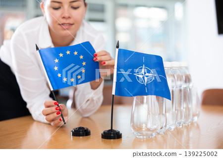 Female secretary places flags of the NATO (OTAN) and European Union flag on the table before negotiations of top political figures Female secretary places flags of the NATO (OTAN) and European Union flag on the table before negotiations of top political figures 139225002