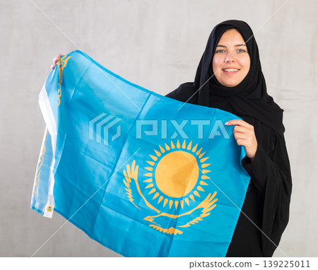 Smiling joyful Muslim woman in traditional black hijab holds flag of Kazakhstan 139225011