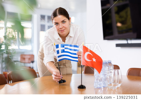 Positive young woman putting flag of Greece on the table with flag of Turkey in conference room Positive young woman putting flag of Greece on the table with flag of Turkey in conference room 139225031