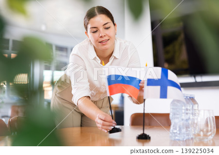 Positive young woman putting flag of Russia on the table with flag of Finland in conference room Positive young woman putting flag of Russia on the table with flag of Finland in conference room 139225034